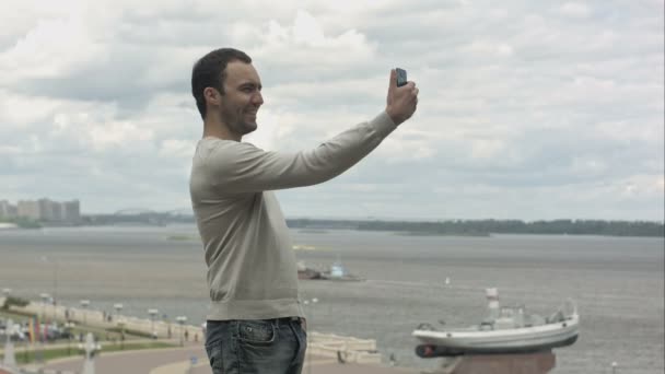 Giovane fa selfie con il telefono cellulare vicino al fiume il giorno nuvoloso — Video Stock