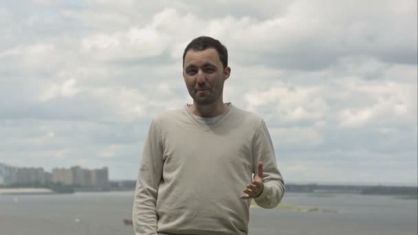 Joven hombre expresivo de pie junto al río, conversando y haciendo gestos con los brazos y las manos a la cámara, al aire libre . — Vídeos de Stock