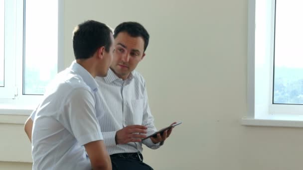 Два бизнес-руководителя сидят в светлом офисе и ищут информацию вместе, делясь экраном цифрового планшета — стоковое видео