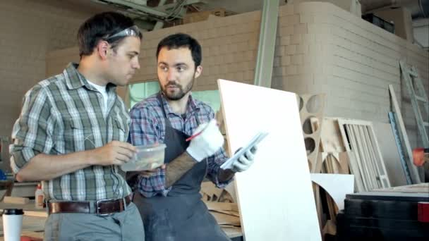 Dois carpinteiros discutindo o trabalho um deles comendo e outro escrevendo em caderno . — Vídeo de Stock