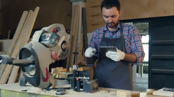 Carpenter pracuje na tabletu poblíž elektrická řetězová pila v dílně. — Stock video