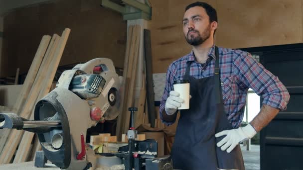 Der Tischler trinkt Kaffee und denkt über etwas in der Werkstatt in der Nähe der elektrischen Säge nach. — Stockvideo