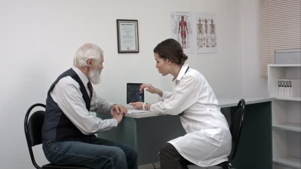 Femme médecin parlant avec le patient âgé dans le bureau et lui montrant la tablette avec IRM . — Video