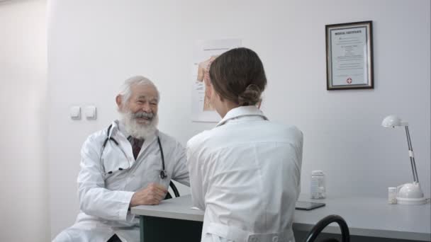 Un médico de alto rango en un escritorio en un consultorio sonriendo y hablando con una doctora . — Vídeos de Stock