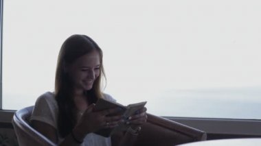 Young beautiful girl sits in a cafe with panoramic views of atlantic ocean. Morning with book reading, thinking