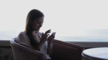 young beautiful girl sits in a cafe with panoramic views of atlantic ocean. Useing smart phone, smiling
