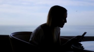 gadget kullanarak ve Atlantik Okyanusu'nun panoramik manzaralı bir kafede oturan genç güzel kız silueti. Sabah, akıllı telefon kullanarak, internet