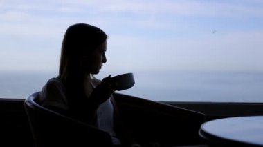 genç güzel kadın siluet atlantik okyanusunun panoramik manzaralı bir kafede oturur. Bir fincan coffe, çay, içme, düşünme, bekletme ile Sabah