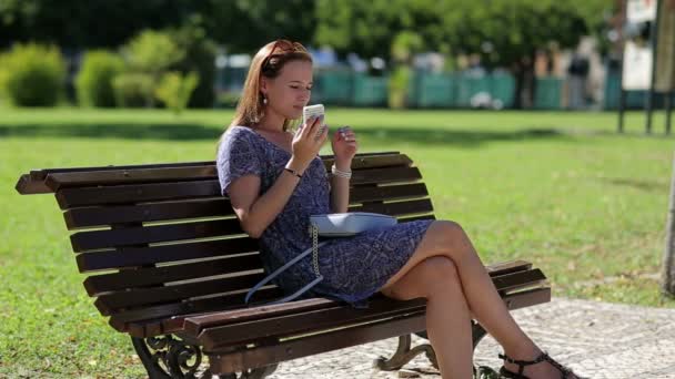 Jovem mulher bonita segurando telefone celular na mão e sentado no banco no parque — Vídeo de Stock