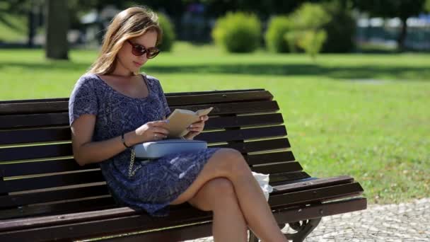 Young woman reading a book and sitting on a bench outside in a park in summer, in sunglasses — Stockvideo