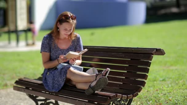 Young woman relaxing, reading a book and sitting on a bench with legs outside in a park in summer, bird in background — 图库视频影像