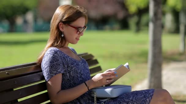 Jeune femme n'aime pas lire un livre et assis sur un banc à l'extérieur dans un parc en été, dans des lunettes de soleil avec sac à main, bain de soleil et se détendre 