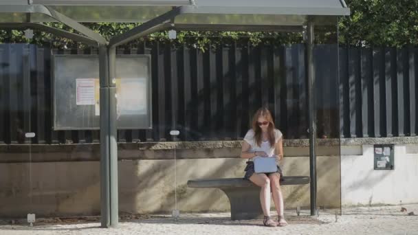 Μοναχικό νεαρό κορίτσι που περιμένει στη στάση του λεωφορείου με έξυπνο τηλέφωνο σε μπλε φούστα και γυαλιά ηλίου, καλοκαίρι — Αρχείο Βίντεο
