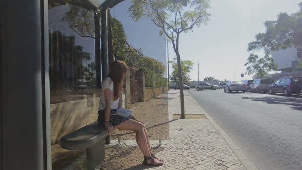 Lisboa, Portugal - Septiembre 2015: Mujer joven y solitaria esperando en la parada de autobús de cristal en falda con teléfono inteligente y bolso, verano — Vídeos de Stock