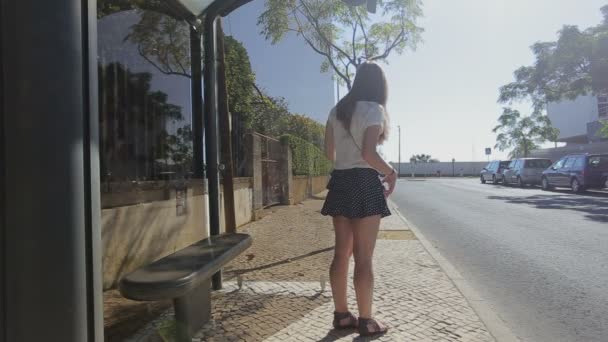 Carcavelos, Portugal - September 2015: Lonely young girl waiting at bus stop with smarphone in blue skirt and sunglasses, summertime, traffic — стоковое видео