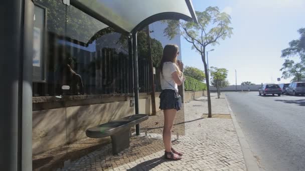 Carcavelos, Portugal - Septiembre 2015: niña esperando sentada en la parada de autobús con teléfono inteligente en falda azul y gafas de sol, verano, y comunicarse — Vídeo de stock