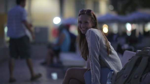 Hermosa chica sentada en un banco en el terraplén por la noche sonriendo y mirando a lo lejos — Vídeos de Stock