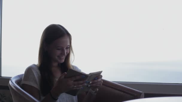 Giovane bella ragazza si siede in un caffè con vista panoramica sull'oceano Atlantico. Mattina con lettura del libro, pensando — Video Stock