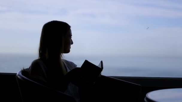 Silhueta de menina bonita jovem senta-se em um café com vistas panorâmicas do oceano atlântico. Ler um livro, passear, pensar, apreciar a vista — Vídeo de Stock