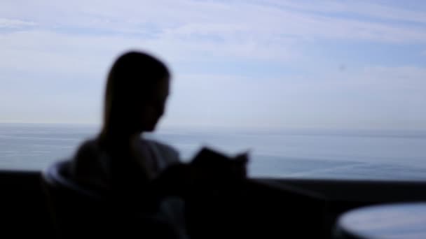 Силуэт молодой красивой девушки сидит в кафе с панорамным видом на Атлантический океан. Утро с книгой, блокнот, чтение, размышления — стоковое видео