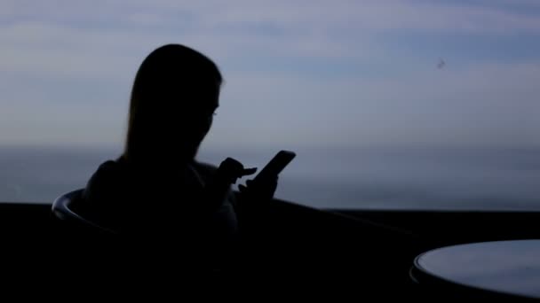 Σιλουέτα του νεαρού κοριτσιού κάθεται σε ένα καφέ με πανοραμικό παράθυρο με θέα στον Ατλαντικό ωκεανό. Χρήσηςέξυπνο τηλέφωνο, συζήτηση — Αρχείο Βίντεο