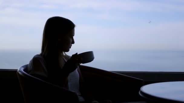 Silhueta de jovem bela mulher senta-se em um café com vistas panorâmicas do oceano atlântico. Manhã com uma xícara de café, chá, beber, pensar, contemplar — Vídeo de Stock