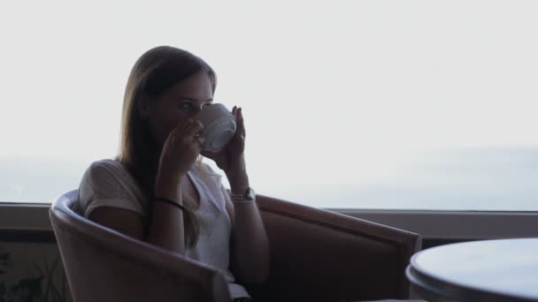Young beautiful girl sits in a cafe with beautiful panoramic views of atlantic ocean. Morning with a cup of coffe, tea — 图库视频影像