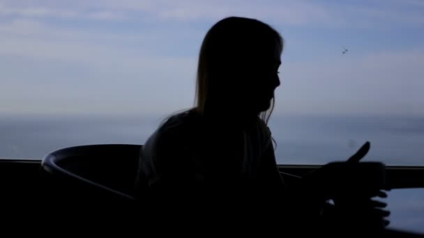 Silueta de joven hermosa niña se sienta en un café con vistas panorámicas del océano atlántico contemplando barco de peces. Mañana con una taza de café, té, libro, bebida, pensamiento — Vídeos de Stock