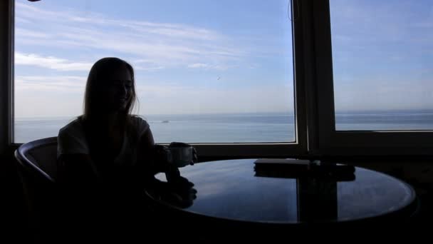 Largo tiro de silueta de joven hermosa niña se sienta en un café con vistas panorámicas del océano atlántico. Mañana con una taza de café, té, libro, bebida, pensamiento — Vídeos de Stock