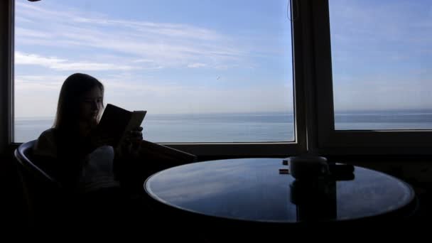 Largo tiro de silueta de tiro largo de joven hermosa niña se sienta en un café con vistas panorámicas del océano atlántico. Mañana con una taza de café, té, libro, bloc de notas, lectura, pensamientos — Vídeos de Stock