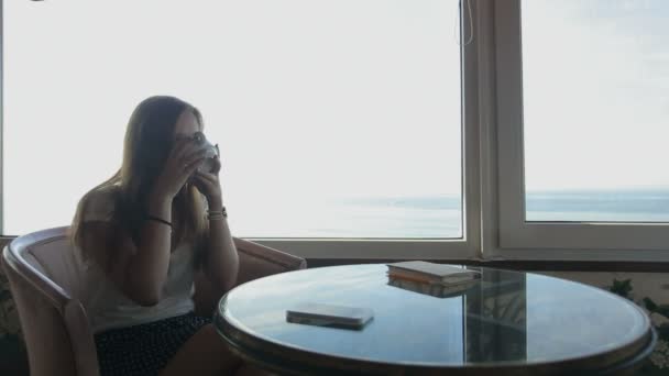 Largo tiro de joven hermosa chica se sienta en un café con vistas panorámicas del océano atlántico. Mañana con una taza de café, té, libro, teléfono inteligente, beber, pensar — Vídeos de Stock