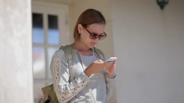 Primer plano de la joven chica bonita en gafas de sol turista en la parada de autobús utilizando el teléfono inteligente con mochila. En el campo. Europa . — Vídeos de Stock