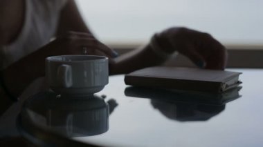 genç güzel kız yakın elleri panoramik manzaralı bir kafede oturur. Sabah bir fincan coffe, çay, kitap, not defteri, okuma, wrighting, içme, iletişim, smrt telefon kullanımı