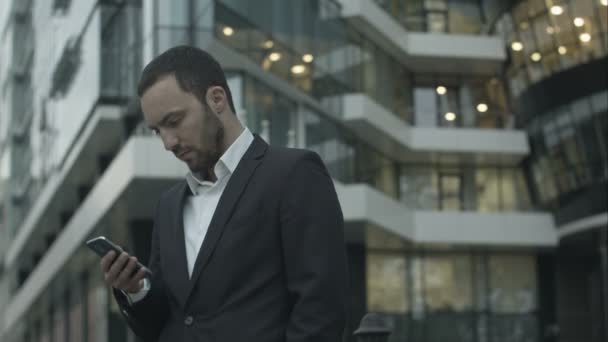 Молодой бизнесмен читает текстовое сообщение, стоящее возле офиса — стоковое видео