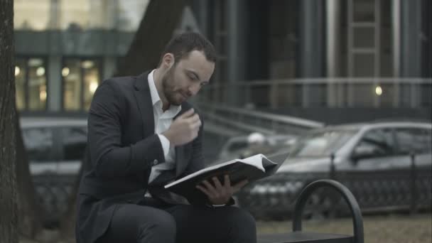 Man i kostym läsa affärs plan med intresse, sitter på en bänk i parken och — Stockvideo