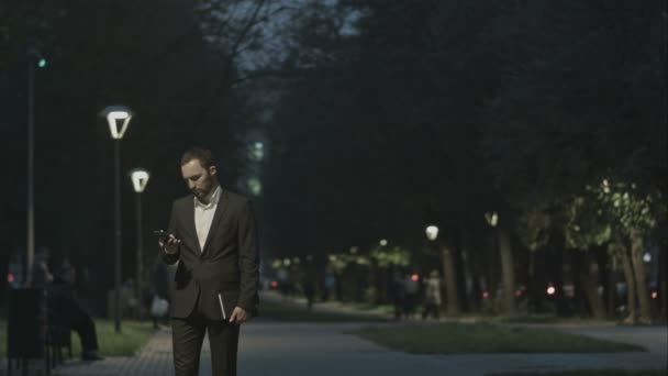 Portréja csinos felnőtt üzletember küld egy szöveges üzenetet állva a város utcán, üzletember üzenet olvasás, stílusos barna mobiltelefon segítségével a szabadban este, Park — Stock videók