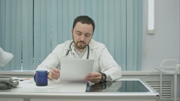 Stanco dal lavoro, medico barbuto bere da tazza e continuare a woking con documenti e radiografie — Video Stock