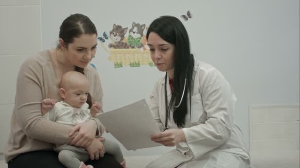 Médico pediatra fêmea mostra alguns papéis para a mulher com bebê pequeno — Vídeo de Stock