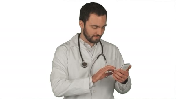 Médico varón médico enviando un mensaje con su teléfono celular mientras sonríe y de pie sobre fondo blanco — Vídeos de Stock