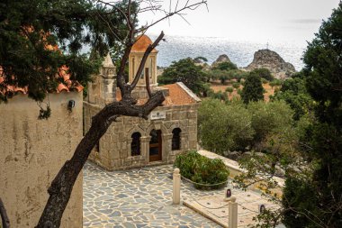 Kıyı koyundaki taş şapel, küçük çan kulesi ile yıpranmış cephe, eğri büğrü zeytin ağacı ön plan,