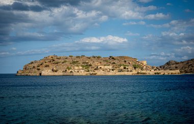 Derin mavi deniz, dağınık bulutlar, durgun su ve sahil savunma kalıntılarına karşı alçak adacık kale silueti.,