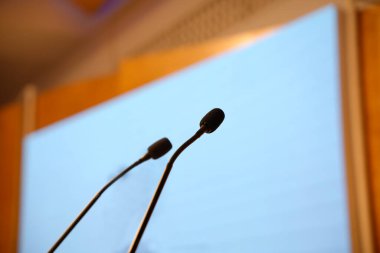 İki kaz boyunlu mikrofon bir konferans odasında kürsüde parlak bir ekranla duruyor.