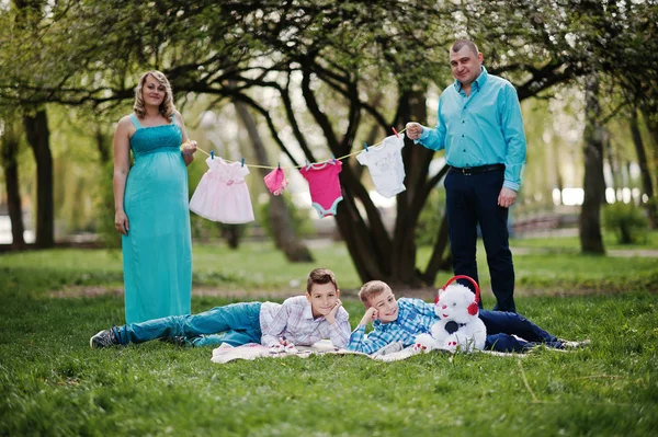 Feliz casal grávida com dois filhos segurando roupas de bebê de meninas — Fotografia de Stock