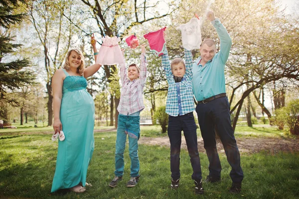 Feliz casal grávida com dois filhos segurando roupas de bebê de meninas — Fotografia de Stock
