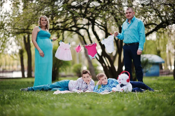 Feliz casal grávida com dois filhos segurando roupas de bebê de meninas — Fotografia de Stock