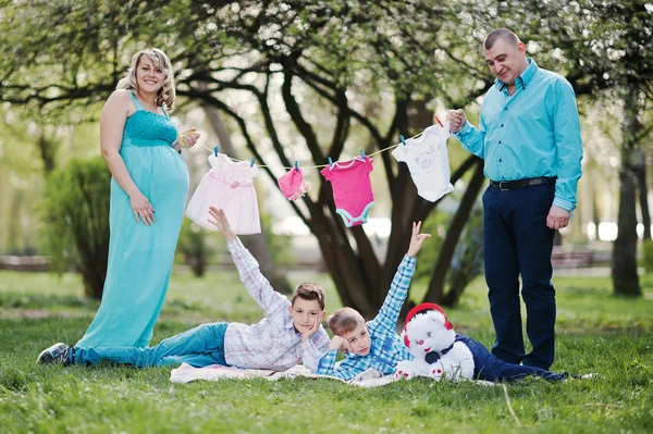 Feliz casal grávida com dois filhos segurando roupas de bebê de meninas — Fotografia de Stock