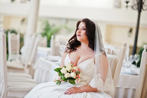 Big breasts brunette bride with wedding bouquet sitting on chair — Stock Photo, Image