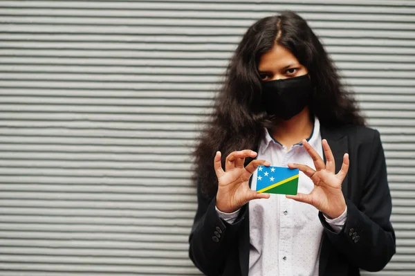 Asian woman at formal wear and black protect face mask hold Solomon Islands flag at hand against gray background. Coronavirus at country concept.