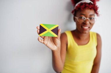 Afro saçlı Afrikalı bir kadın, sarı atlet ve gözlük takıyor, beyaz arka planda Jamaika bayrağı taşıyor..