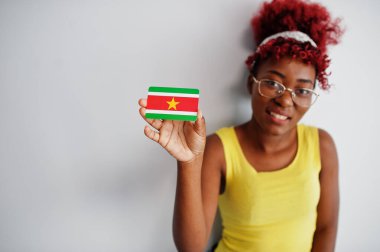 Afro saçlı Afrikalı bir kadın, sarı atlet ve gözlük takıyor, Surinam bayrağını beyaz arka planda tek başına tutuyor..